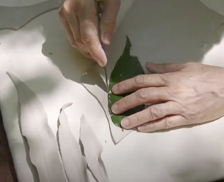 Artist traces a leaf with a blade on a thin layer of clay beside sculpted clay leaves.
