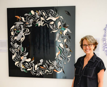 Artist Maria Kraatz standing next to her artwork in a gallery. The artwork is made of over one hundred ceramic leaves, swirls, gumnuts and fish (all of different colours) laid in a broad circle over black glass.