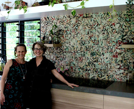 An artist and a house owner stand in front of a splashback of hand-made custom ceramic tiles featuring silohuettes of leaves and flowers.