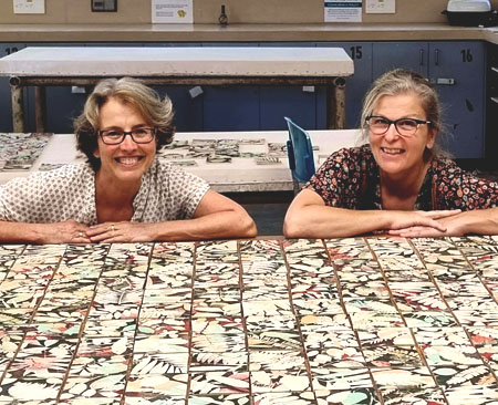 Two artists sit in a studio amongst custom hand-made ceramic tiles featuring silohuettes of leaves and flowers.