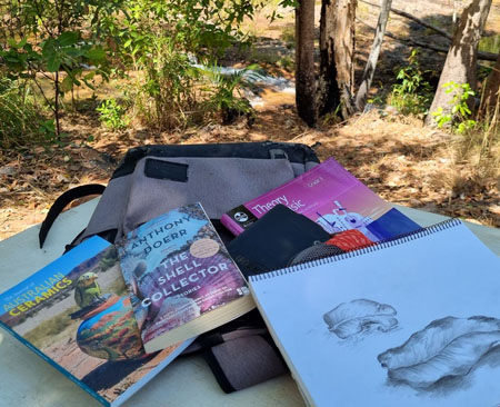 Various books on ceramics and music and a fictional novel and sketch of leaves on top of a backpack in the Australian bush.