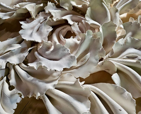 Detail from an artwork called 'Force of Nature' made up of over 80 folded ceramic pieces, ribbons, and leaves, arranged in an outward flow on plywood.