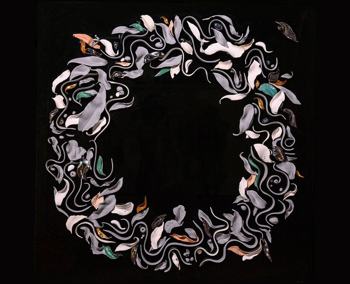 An artwork by Maria Kraatz made up of ceramic leaves, curving lines, stylised fish, pebbles and gum nuts laid in a broad circle on a black glass background.