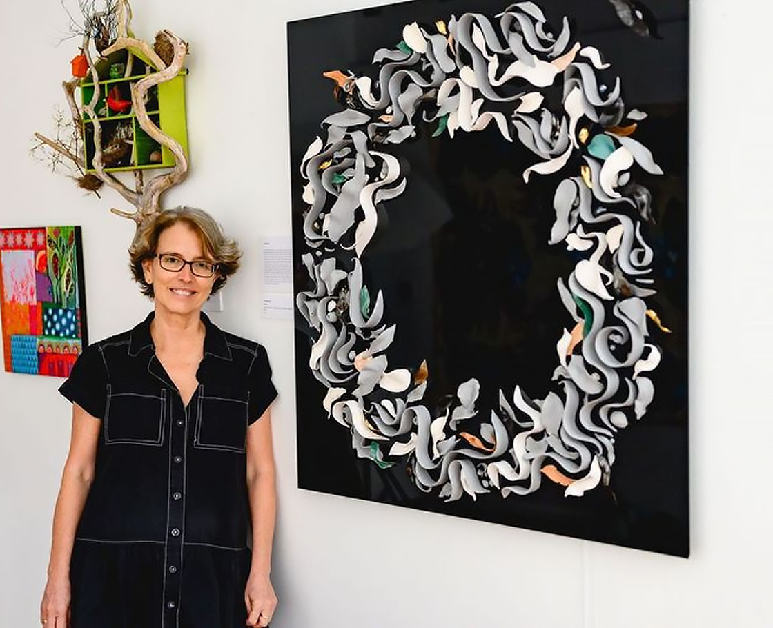 An artist standing next to her artwork in a gallery. The artwork is made of over one hundred ceramic leaves, swirls, gumnuts and fish (all of different colours) laid in a broad circle over black glass.