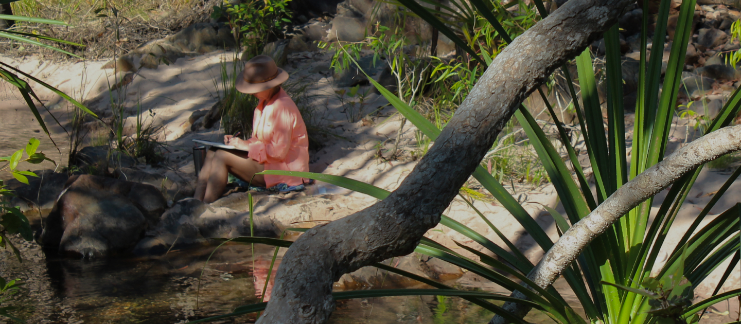 Maria in hat and long sleeved orange shirt sitting on the sand at the edge of a creek and writing in a book.